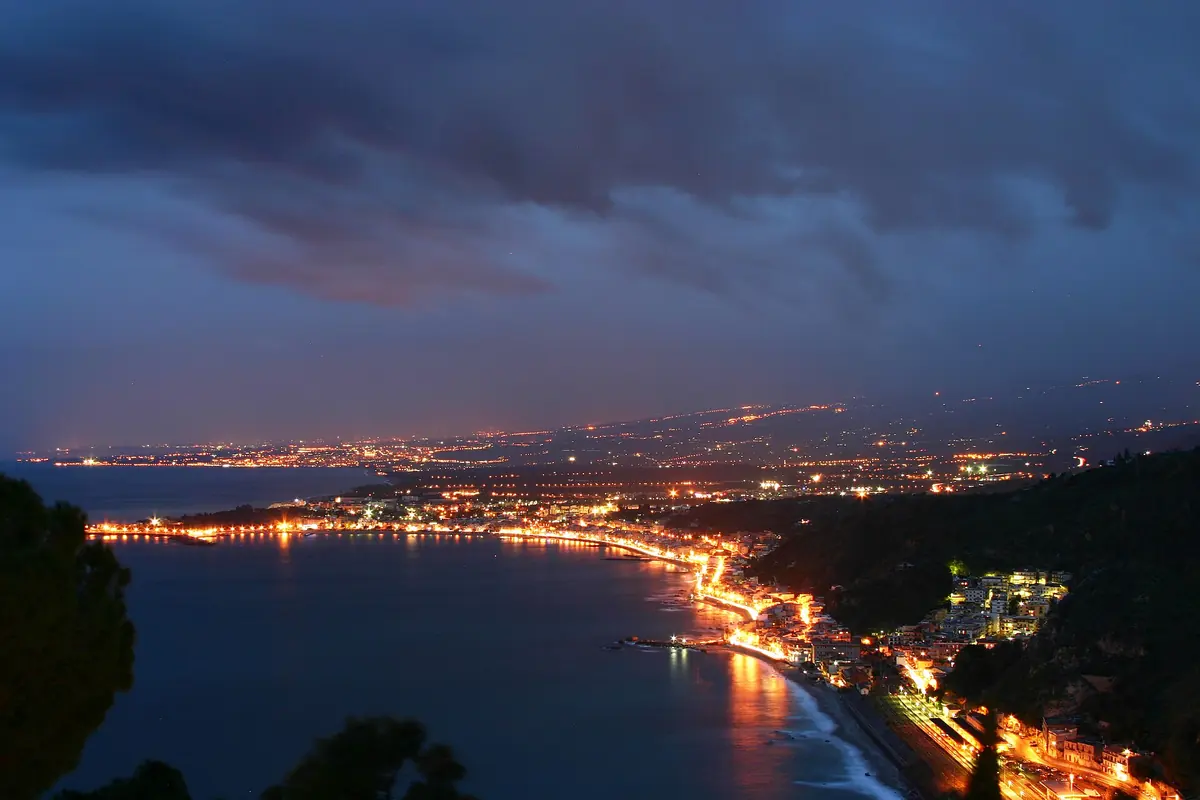 The lights of the coast around the Gulf of Naxos at night, seen from a Taormina hillside under a dark, cloudy sky, with a bright line of road along the shore.