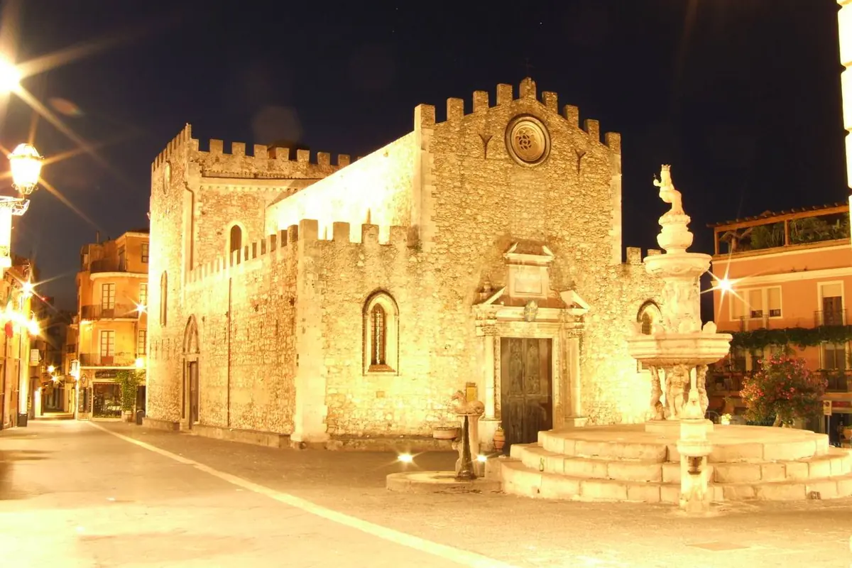 The battlemented front of Taormina Cathedral lit gold at night, with the baroque fountain of Piazza Duomo to its right.