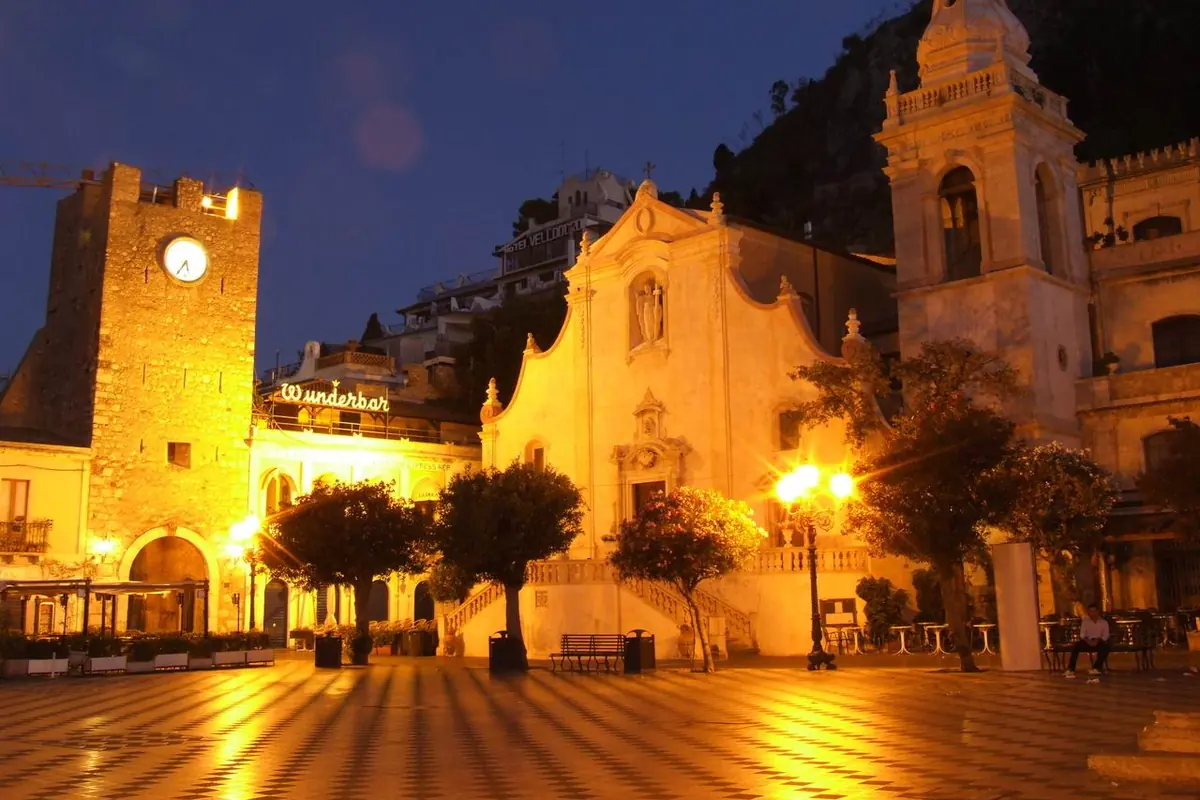 Piazza IX Aprile in Taormina at night, with the lit clock tower on the left, the golden front of the church of San Giuseppe and a bell tower to its right above a chequered square.