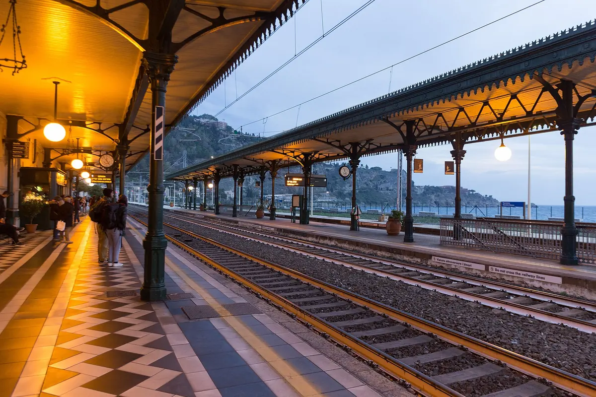The platforms of Taormina-Giardini station under a long iron canopy, with a chequered tiled floor, lamps and the hills above the line.